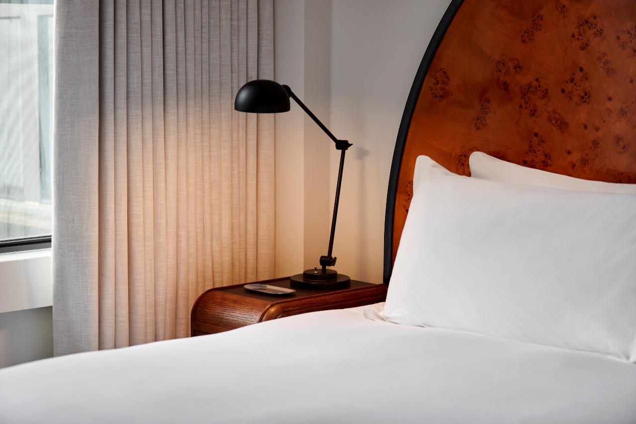 A close-up of a hotel bed with a wooden nightstand, black reading lamp, and neutral-toned curtains at Lyle Washington DC.
