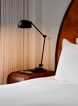 A modern hotel room at Lyle DC with a curved wooden headboard, white bedding, a bedside table, and a black adjustable lamp.