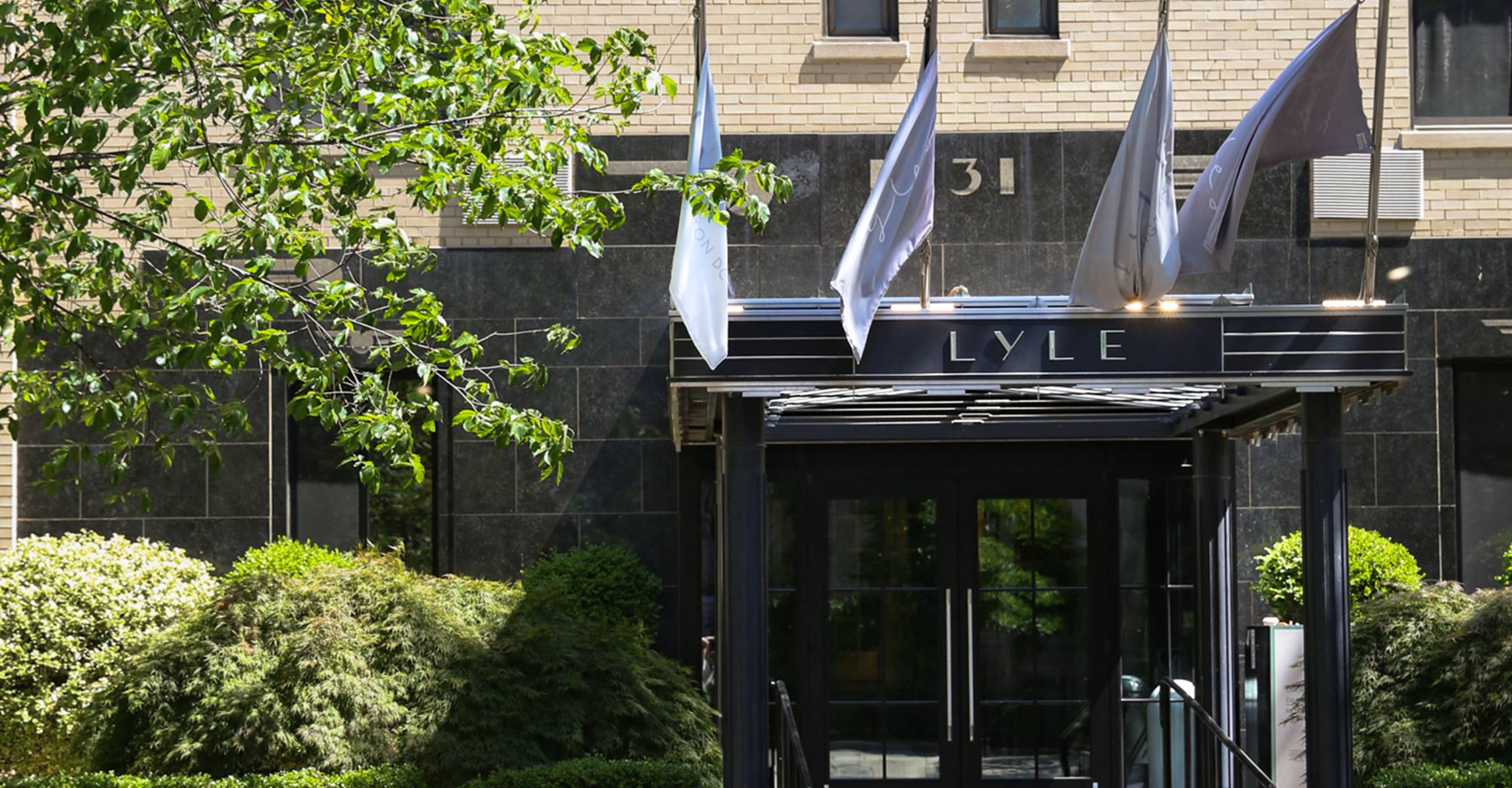 A modern hotel entrance with a black awning, glass doors, and flags, set amidst landscaped gardens and a stone facade.