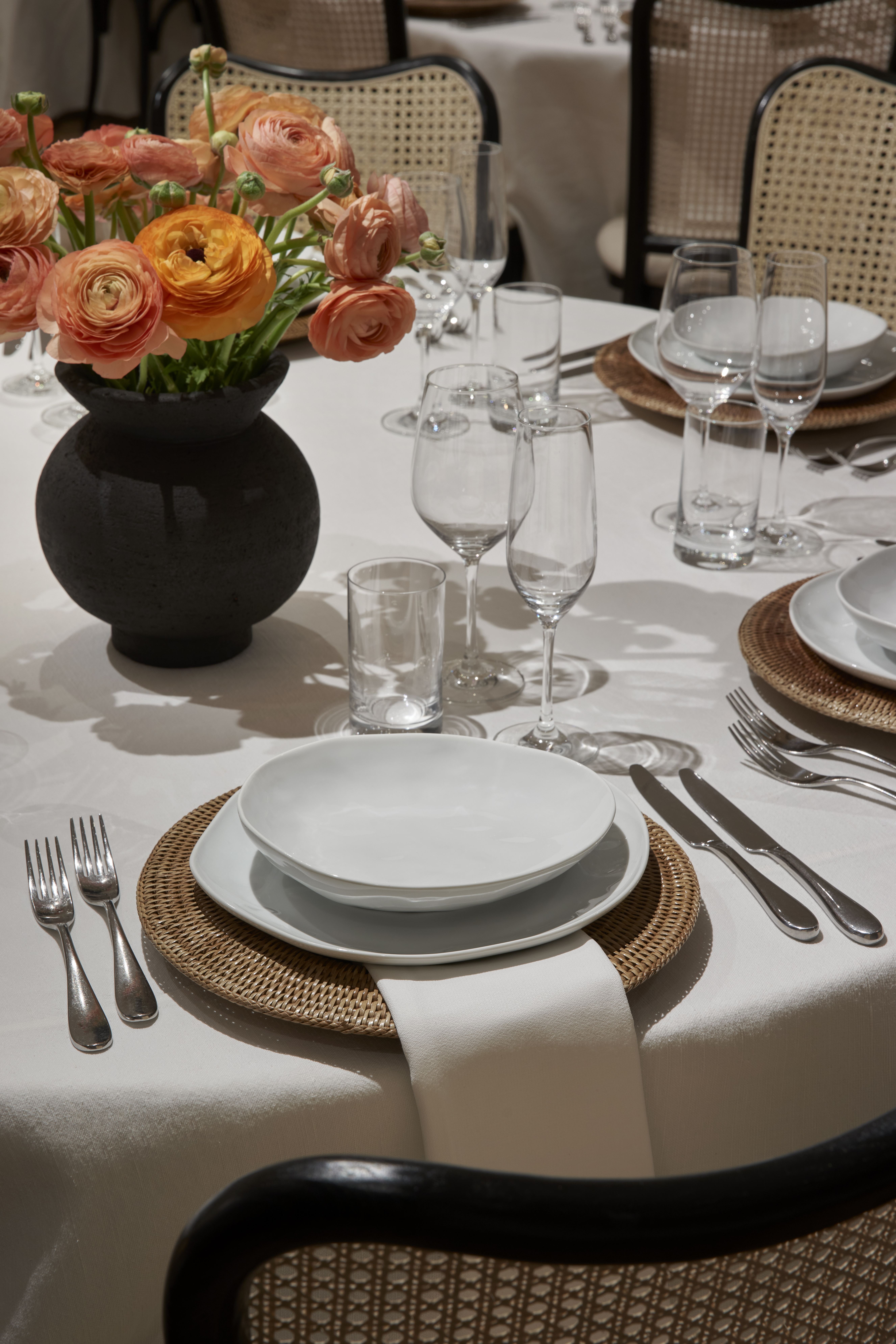 A formal dining table set with white linens, fine china, and glassware, adorned with a floral arrangement in a black vase.