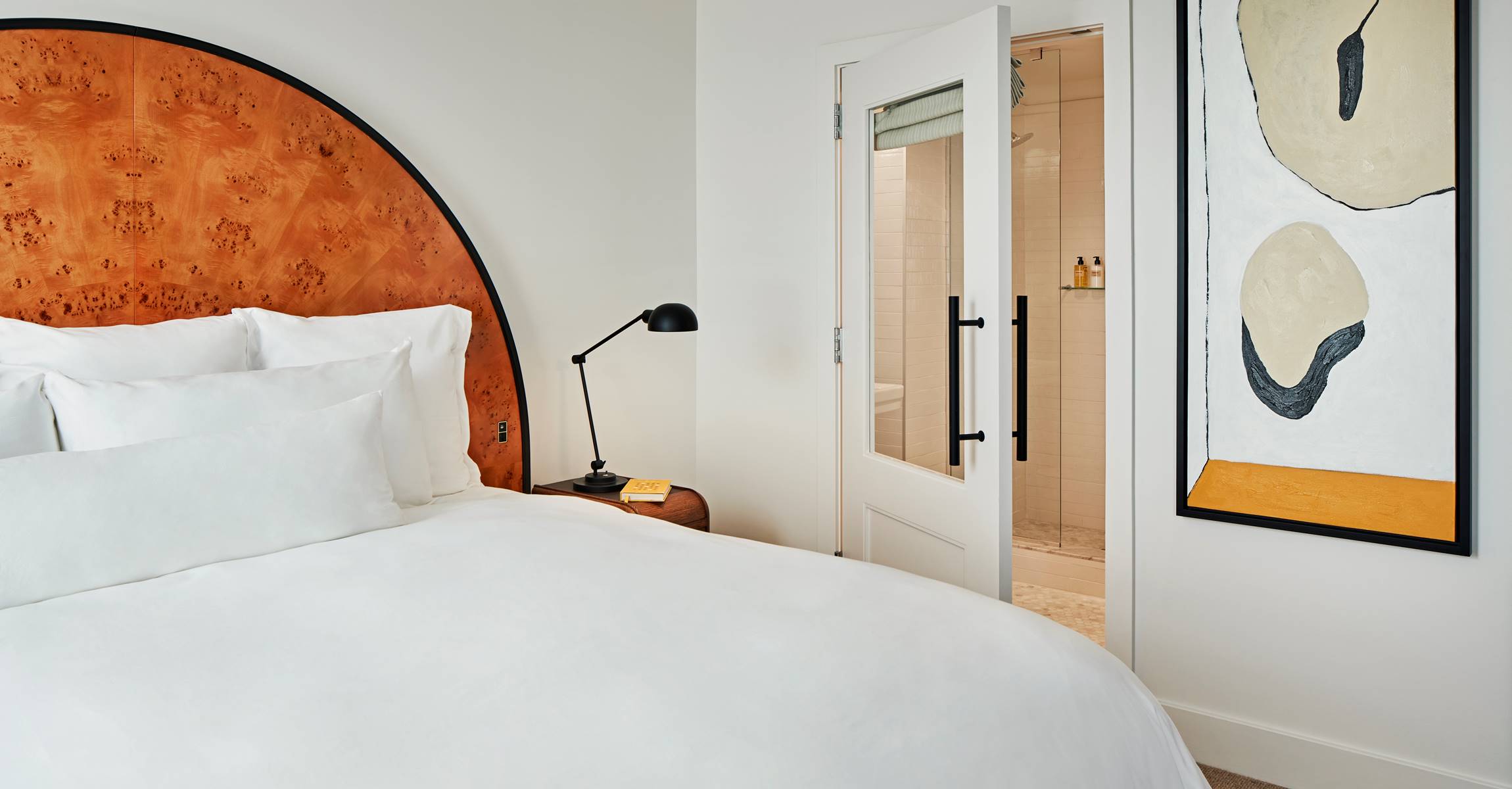 A hotel room with a large bed, wooden headboard, and modern lamp, with a doorway leading to a bathroom with a shower.