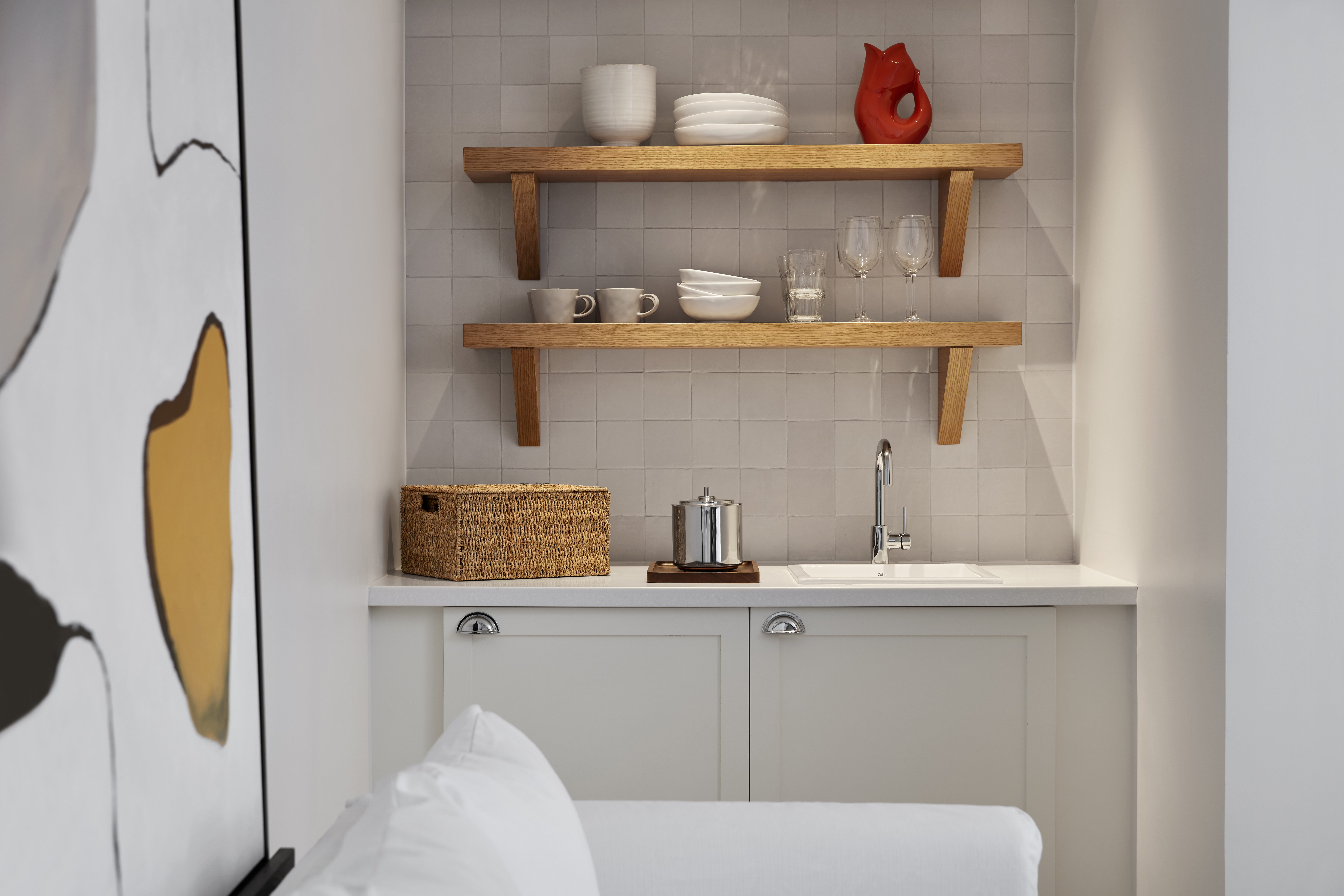 A minimalist kitchenette with white cabinets, open wooden shelving with dishes and glassware, and a woven basket for storage.