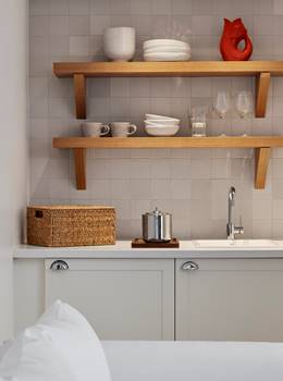A minimalist kitchenette with white cabinets, open wooden shelving with dishes and glassware, and a woven basket for storage.